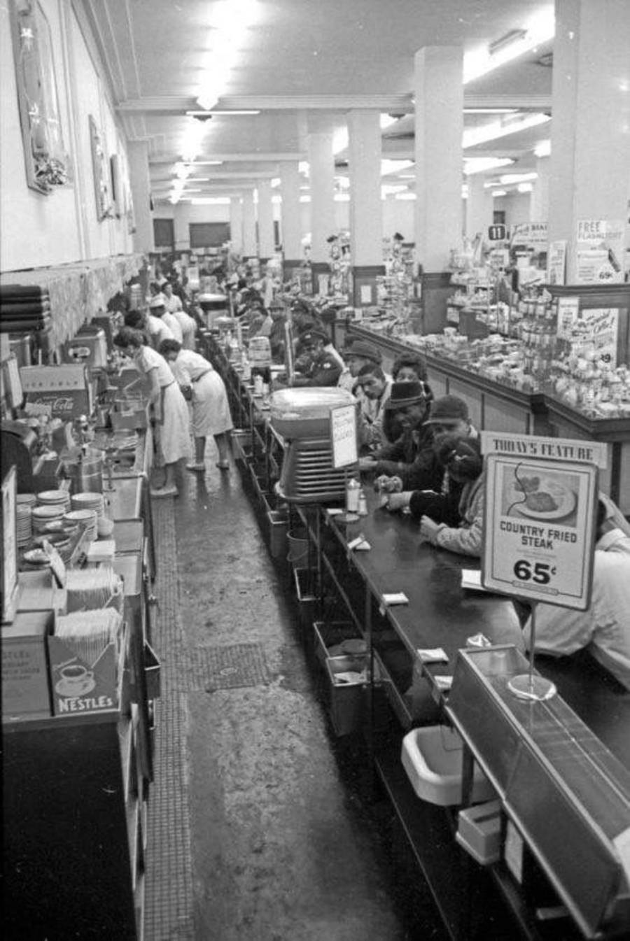 Black-and-white photo of a busy lunch counter in a store. People sit on stools eating, while workers in white uniforms serve food. Signs advertise specials, including "Country Fried Steak 65¢." Shelves of grocery items line the background.
