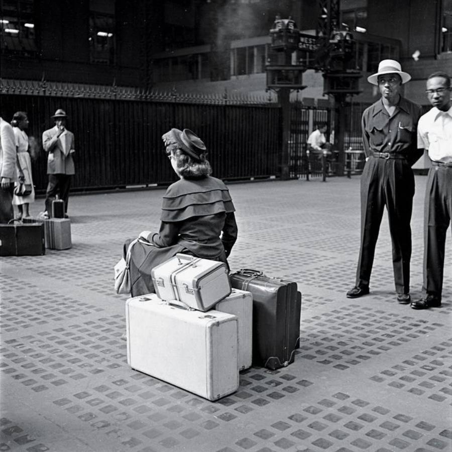 A woman sits on a suitcase among several bags at a train station, while three men stand nearby and another man takes a photo in the background. The scene appears to be from the mid-20th century.