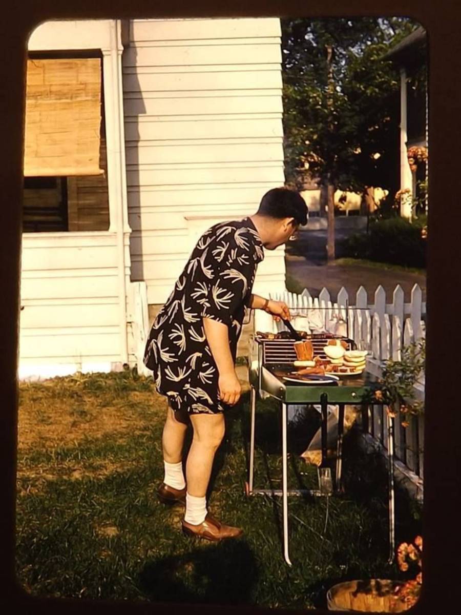 A man wearing shorts and a patterned shirt grills food in a sunny backyard next to a white house and picket fence. Plates and bowls sit on the grill table.