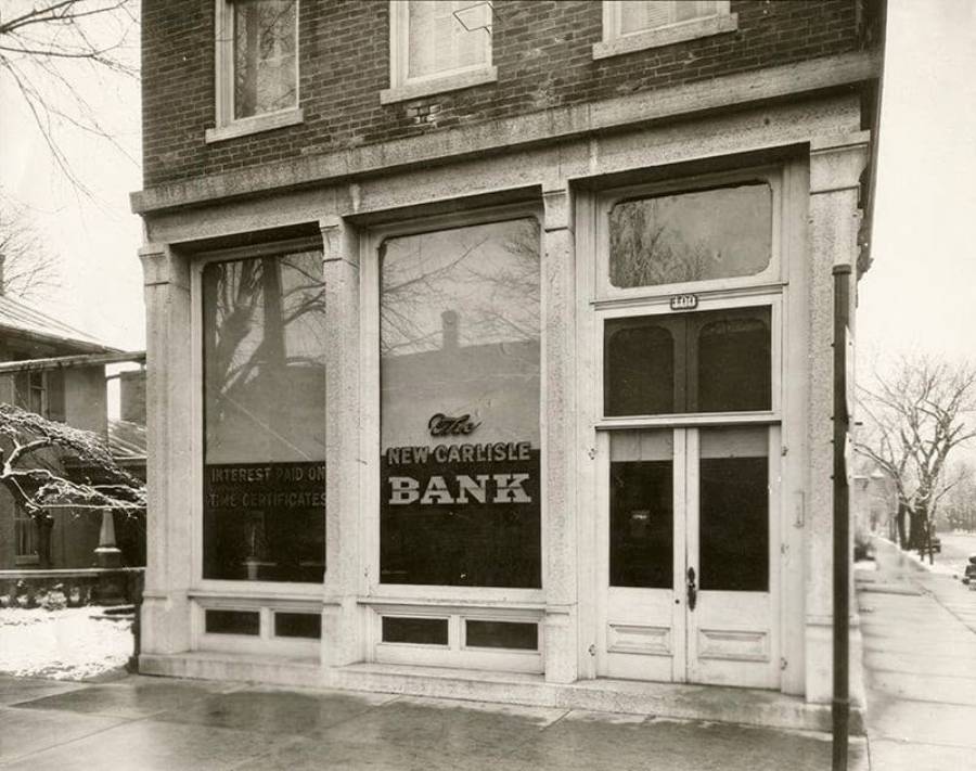 Black-and-white photo of an old-fashioned brick bank building. Large windows display “New Carlisle Bank.” Leafless trees and a sidewalk are visible outside. The building has a tall door and simple architectural details.