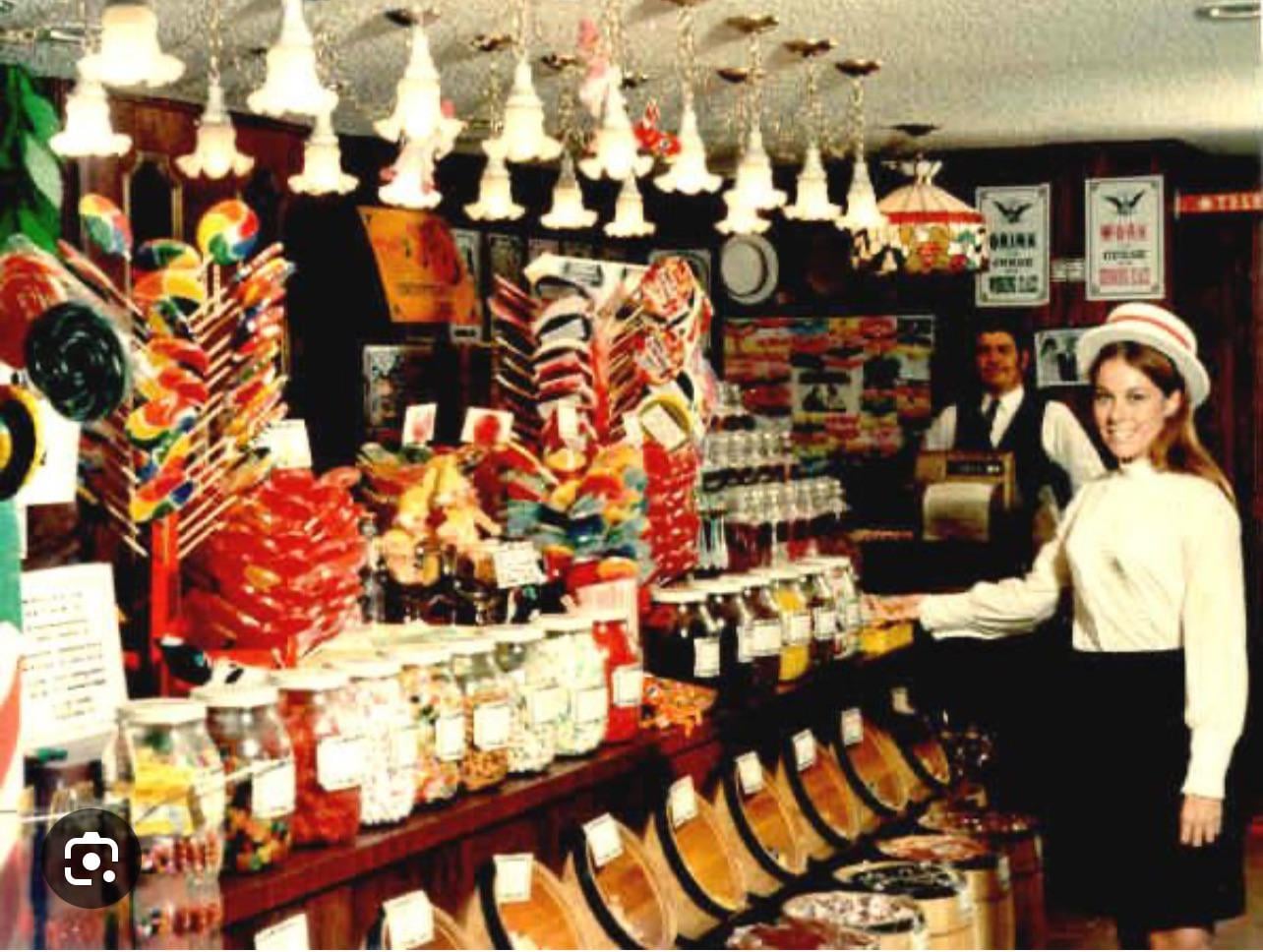 A woman in vintage attire stands in a colorful candy shop, smiling, surrounded by jars of sweets, lollipops, and candies. A man in old-fashioned clothes stands behind the counter.