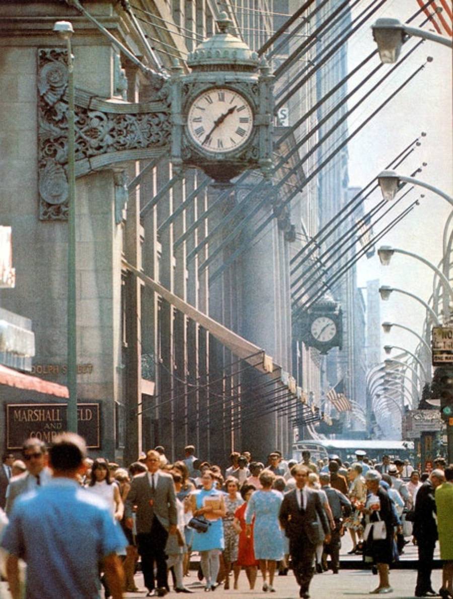 A busy city sidewalk in front of a department store with ornate clocks, crowded with people walking on a sunny day, tall buildings and streetlights lining the street.