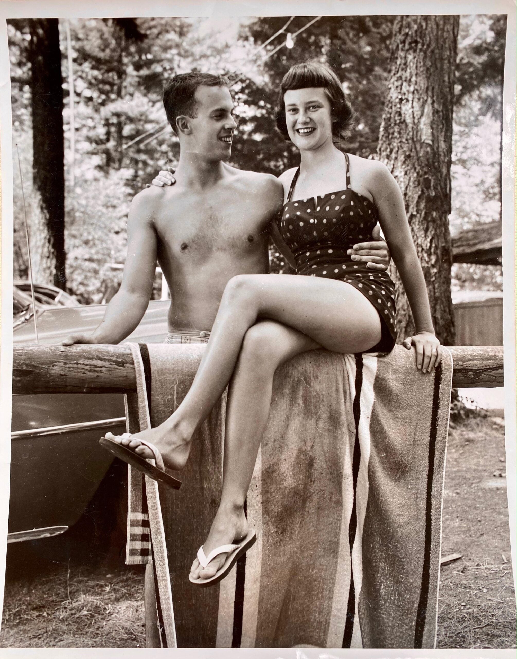 A young man and woman smile while posing outdoors. The man is shirtless and the woman, in a polka-dot swimsuit, sits on a towel-draped fence with her legs crossed. Trees and blurred background suggest a summer setting.