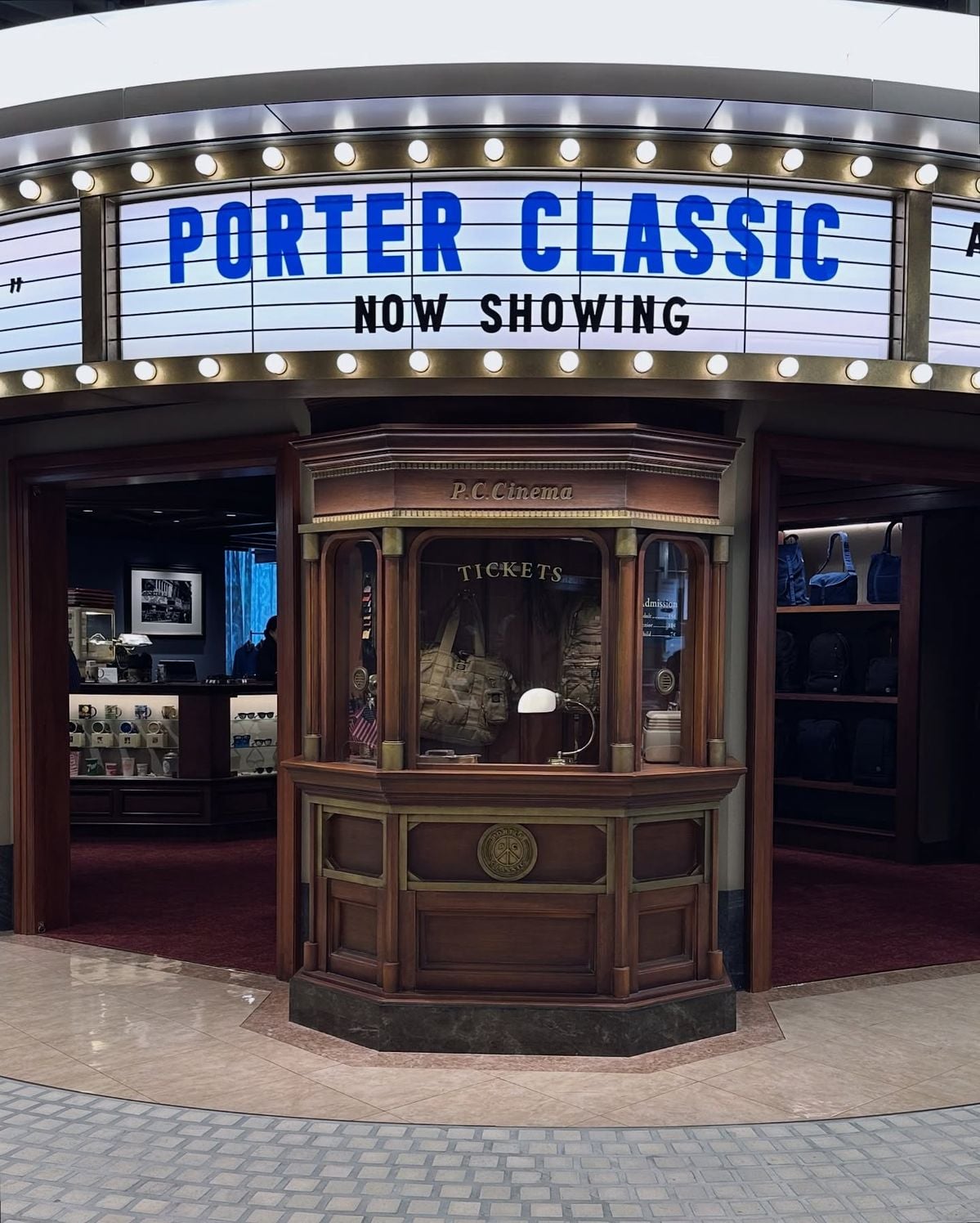 A vintage-style ticket booth sits in front of a shop with a marquee sign above reading “PORTER CLASSIC NOW SHOWING.” The shop interior features displays and shelves with bags and accessories.