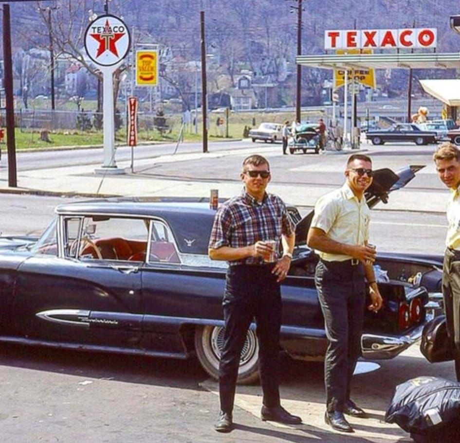 Three men stand beside a classic black car at a Texaco gas station in the 1960s, smiling at the camera. Other cars, trees, and hills are visible in the background.