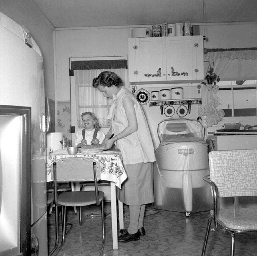 Two women are in a vintage kitchen. One woman sits at a table smiling, while the other stands and prepares food. The room features retro appliances, patterned curtains, and a cabinet with decorative plates.