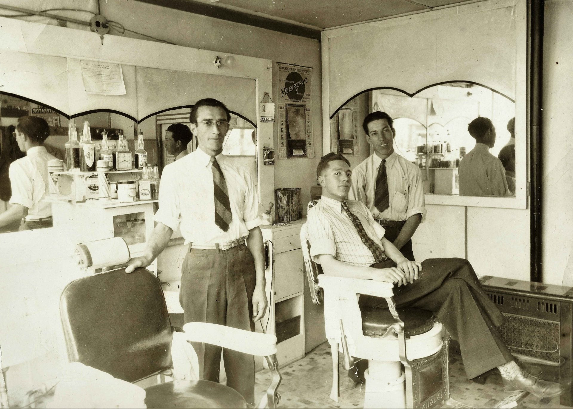 Three men in dress shirts and ties are inside a vintage barbershop; one is seated in a barber chair while the other two stand nearby. Shelves and mirrors with reflections line the background.