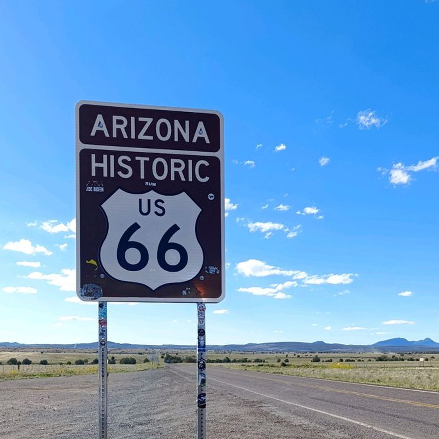 A brown and white road sign reads "Arizona Historic US 66" beside an empty highway under a bright blue sky with scattered clouds and distant mountains.