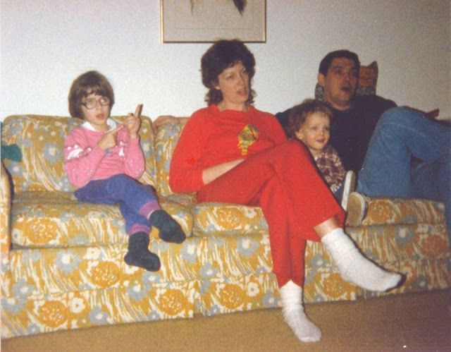 Four people sit on a yellow patterned couch. A young girl in glasses sits at one end, a woman in red is in the center, with a young child on her lap, and a man sits on the other end, all appearing relaxed in a living room.