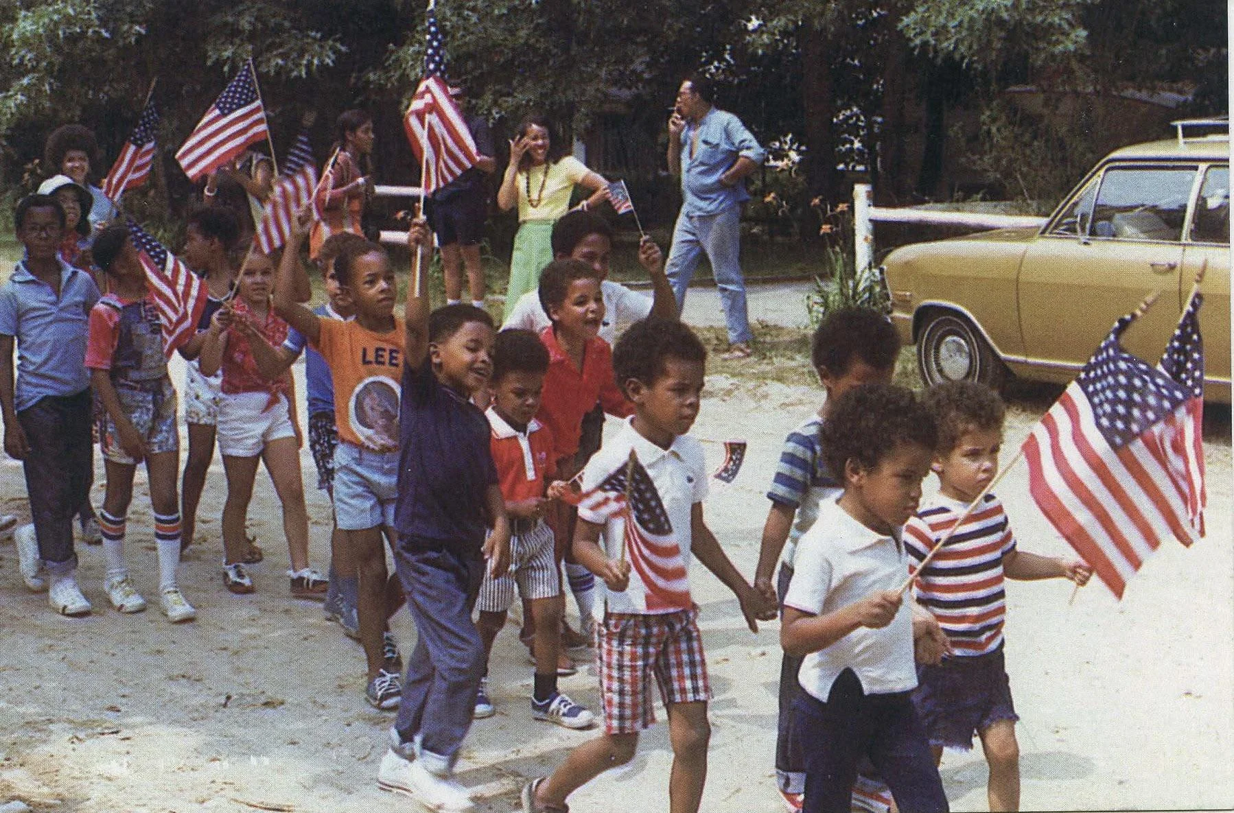 Children march outdoors waving American flags, led by adults. Most are wearing summer clothes with red, white, and blue colors. A tan car is parked nearby, and trees are in the background. The scene appears festive and patriotic.