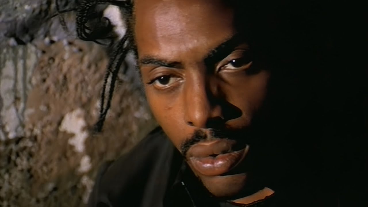 A close-up of a man with braided hair and a mustache, looking down thoughtfully. Part of his face is in shadow, and there is a textured stone wall in the background.