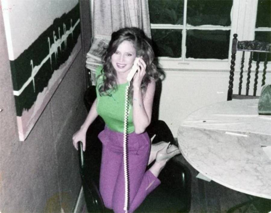 A woman with long hair, wearing a green top and purple pants, sits on a chair by a table, smiling while talking on a corded phone. There is a painting and some papers in the background.