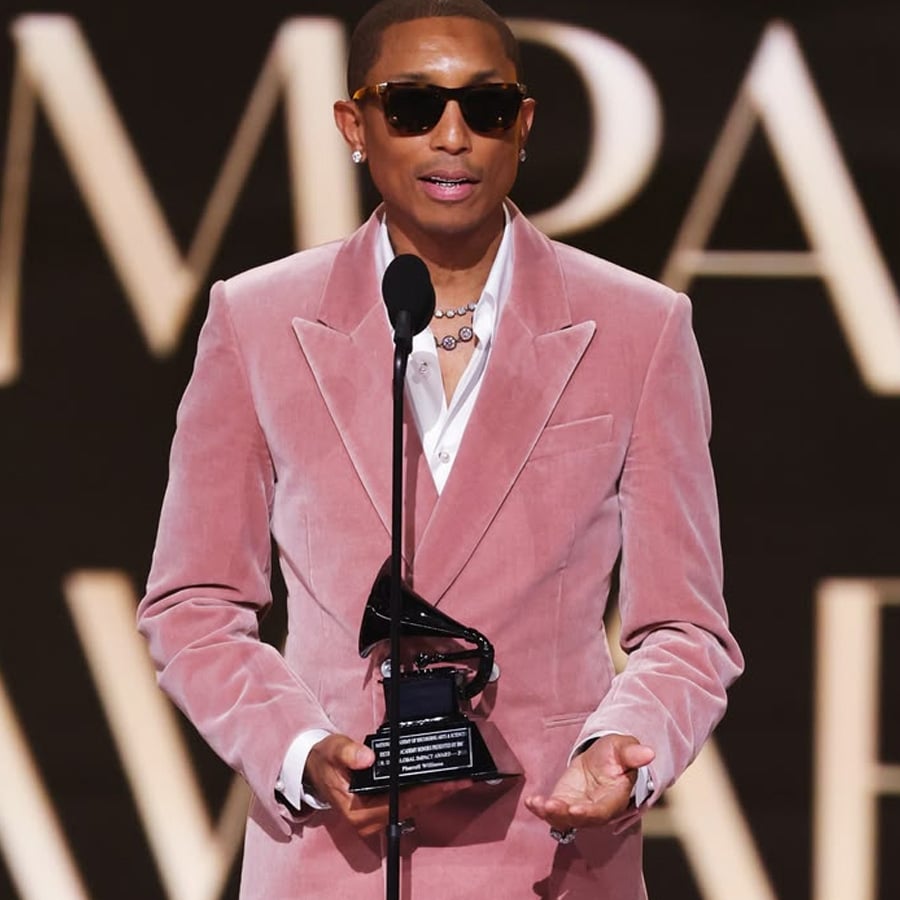 A person in a pink velvet suit and sunglasses holds a Grammy trophy while speaking at a podium on stage.