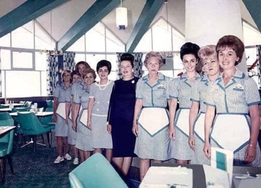A group of women in matching retro waitress uniforms and one in a black dress stand in a line inside a brightly lit vintage diner with teal chairs.