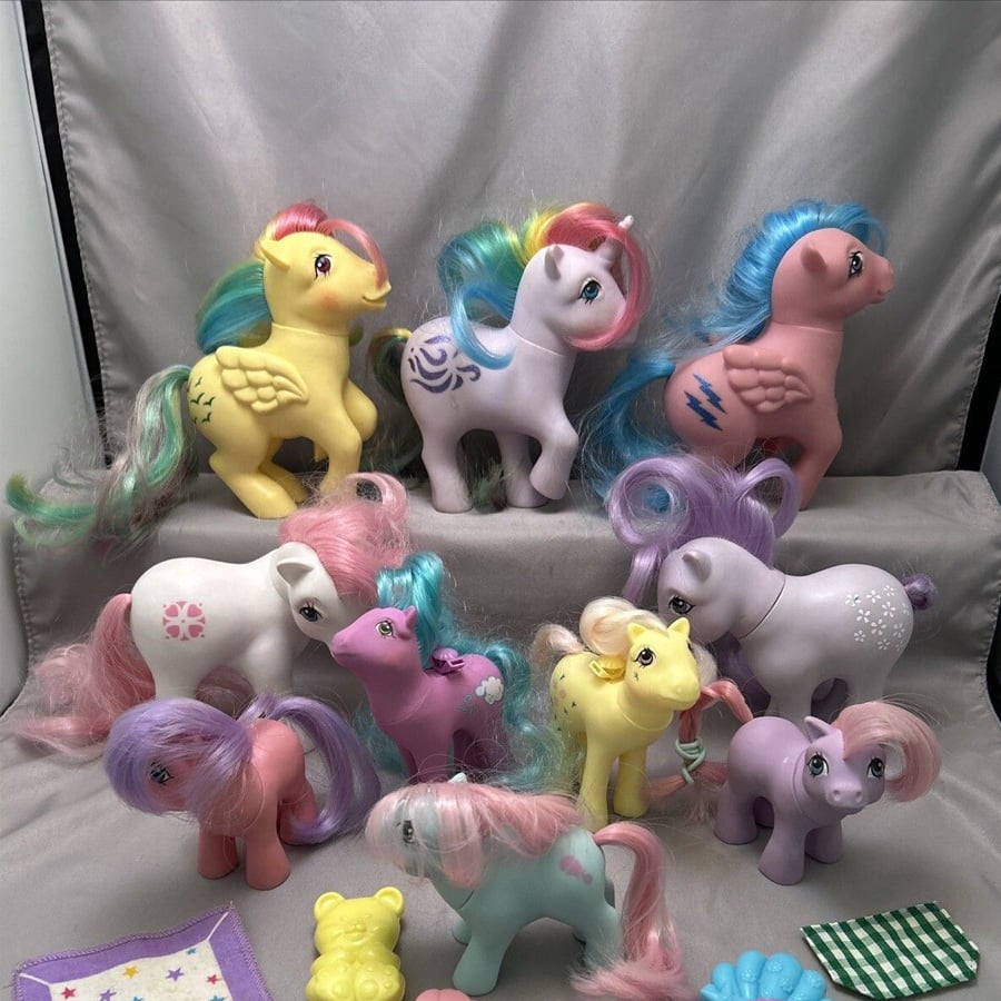 A group of colorful My Little Pony toys with pastel-colored bodies and manes are arranged on gray fabric. Some stand, while others lie down. Small toy accessories and a blanket appear in the foreground.