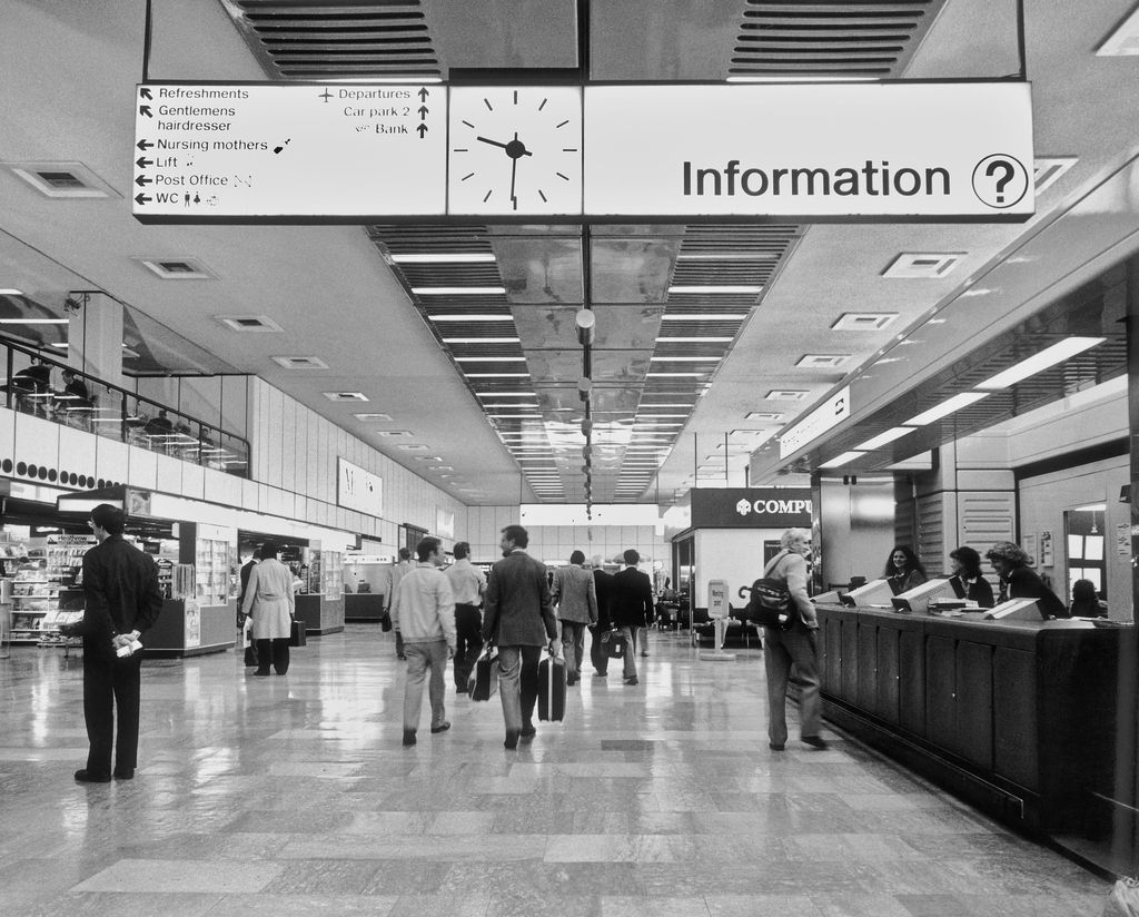 Black and white photo of a busy airport terminal with people walking and an information desk on the right. Overhead signs indicate directions to departures, refreshments, restrooms, nursing mothers' room, and a post office.