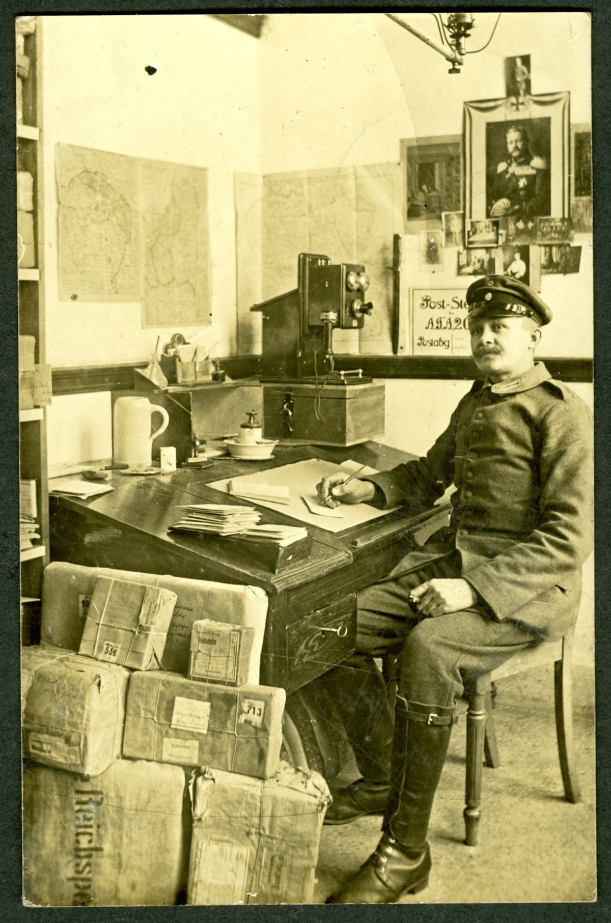 A man in a uniform sits at a desk in an old office with maps on the wall, packages stacked nearby, and a vintage telephone. He appears to be writing or working with papers.