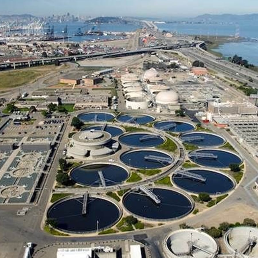 Aerial view of a large wastewater treatment plant with several circular settling tanks, industrial buildings, and surrounding infrastructure near a city waterfront.