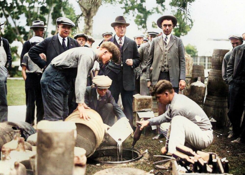 A group of men in early 20th-century attire watch as law enforcement pours liquor from barrels into a drain during a prohibition-era alcohol bust outdoors.