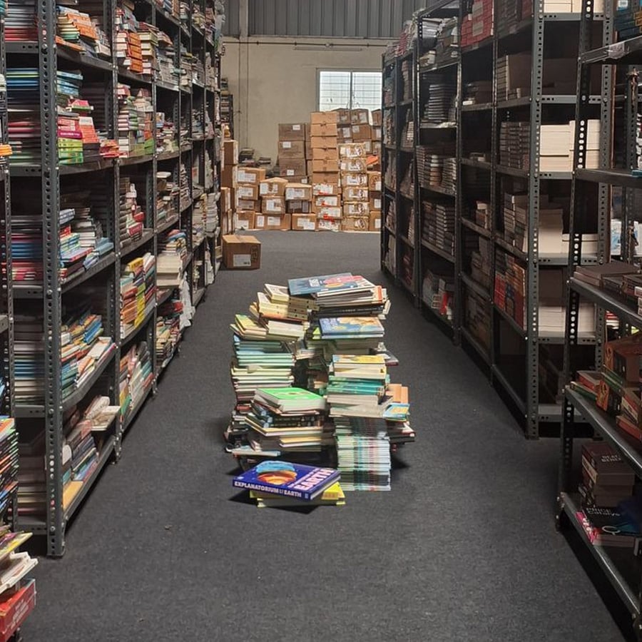 Aisle in a warehouse lined with tall bookshelves, with several stacks of books arranged on the carpeted floor and boxes visible in the distance.
