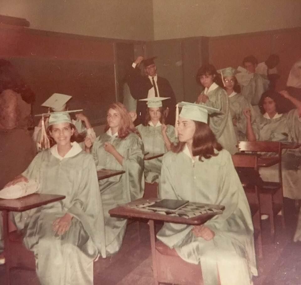 Graduates in caps and gowns sit at desks in a classroom, some smiling. A person in a suit and cap stands at the back, holding papers. The scene appears to be from an earlier decade.