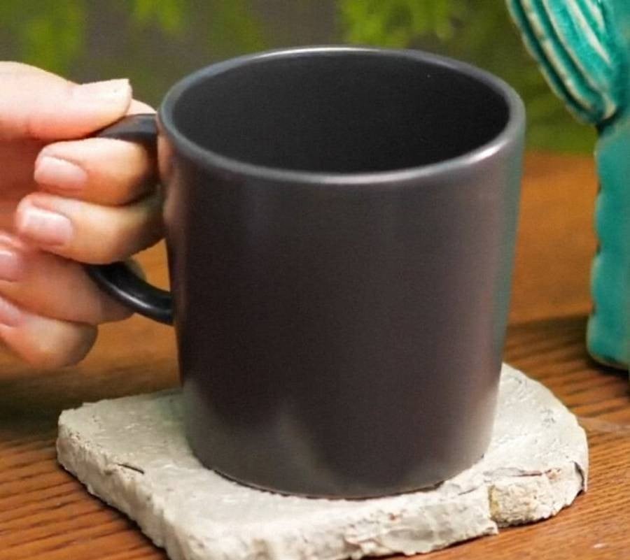A hand holds the handle of a plain black mug resting on a square gray stone coaster, placed on a wooden surface.