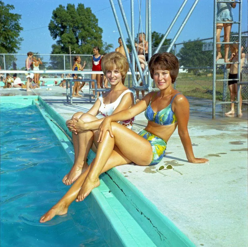 Two women in colorful swimsuits sit by the edge of a pool, smiling at the camera. Other people and children are in the background, enjoying a sunny day at an outdoor pool.