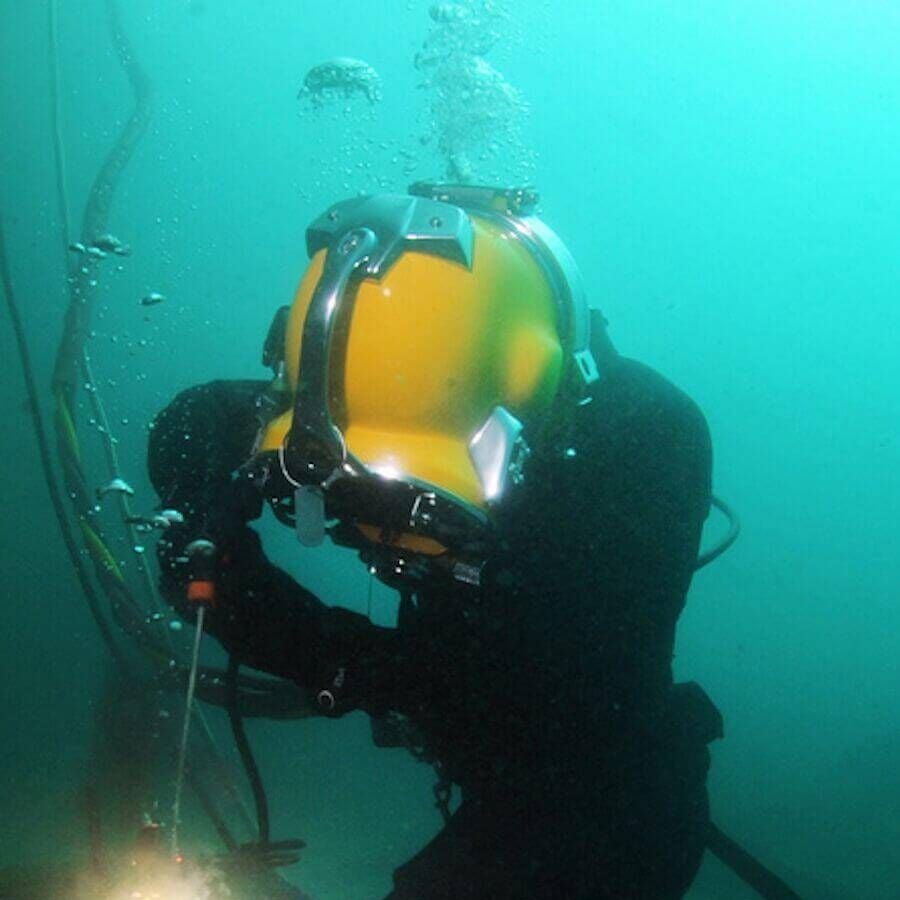 A diver in a yellow helmet and black suit works underwater, holding tools with bubbles rising around them.