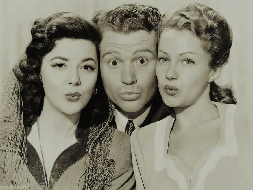 Three people pose closely together, looking at the camera with puckered lips as if blowing kisses. The image is black and white, and they all have neatly styled hair and wear vintage clothing.