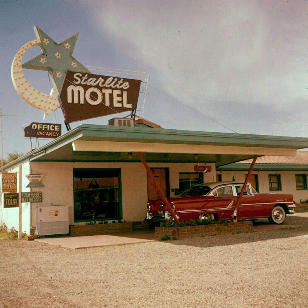 A vintage red car is parked under the slanted awning of the retro Starlite Motel, which features a large star-shaped neon sign. The motel has a classic mid-20th century look.