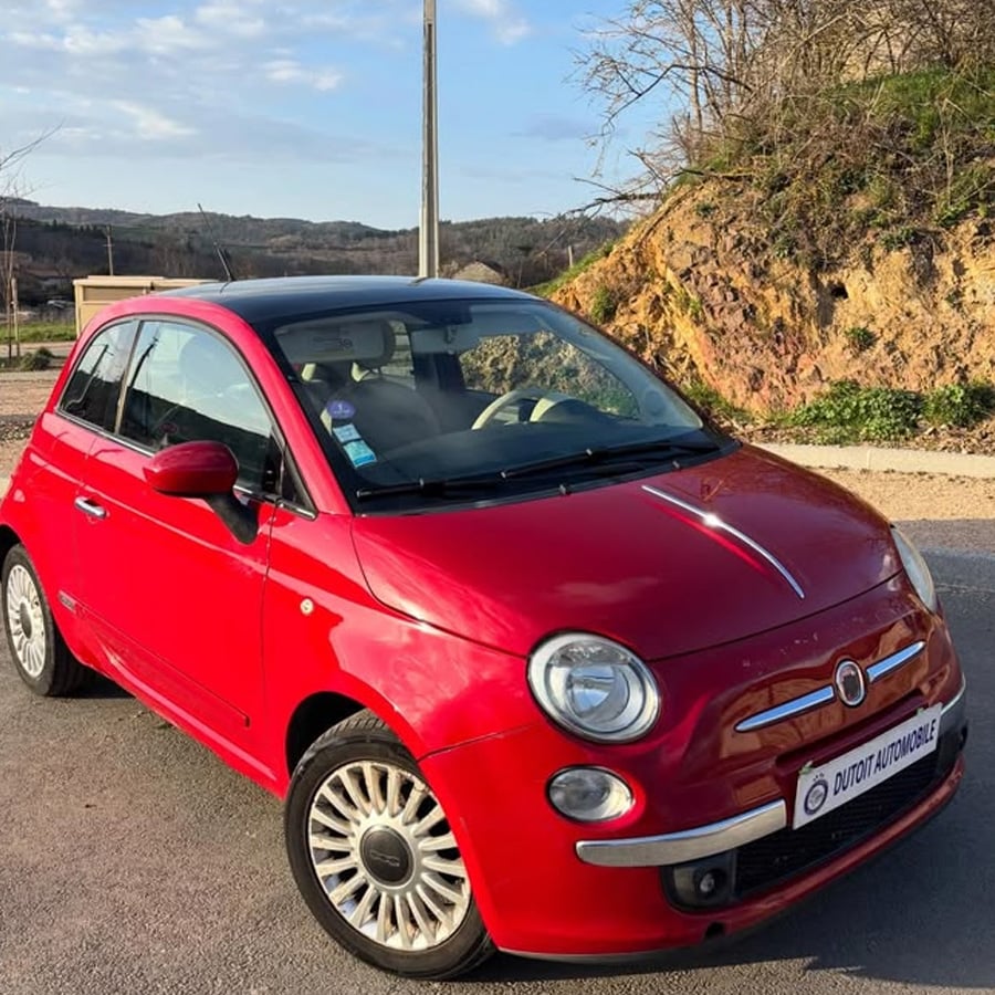 A red Fiat 500 car is parked on a sunlit road, with a rocky hill and trees in the background under a clear sky.