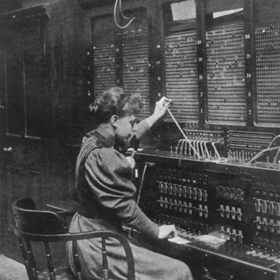 A woman in old-fashioned clothing operates a telephone switchboard, connecting calls using cables and plugs, in a black-and-white historical photo.