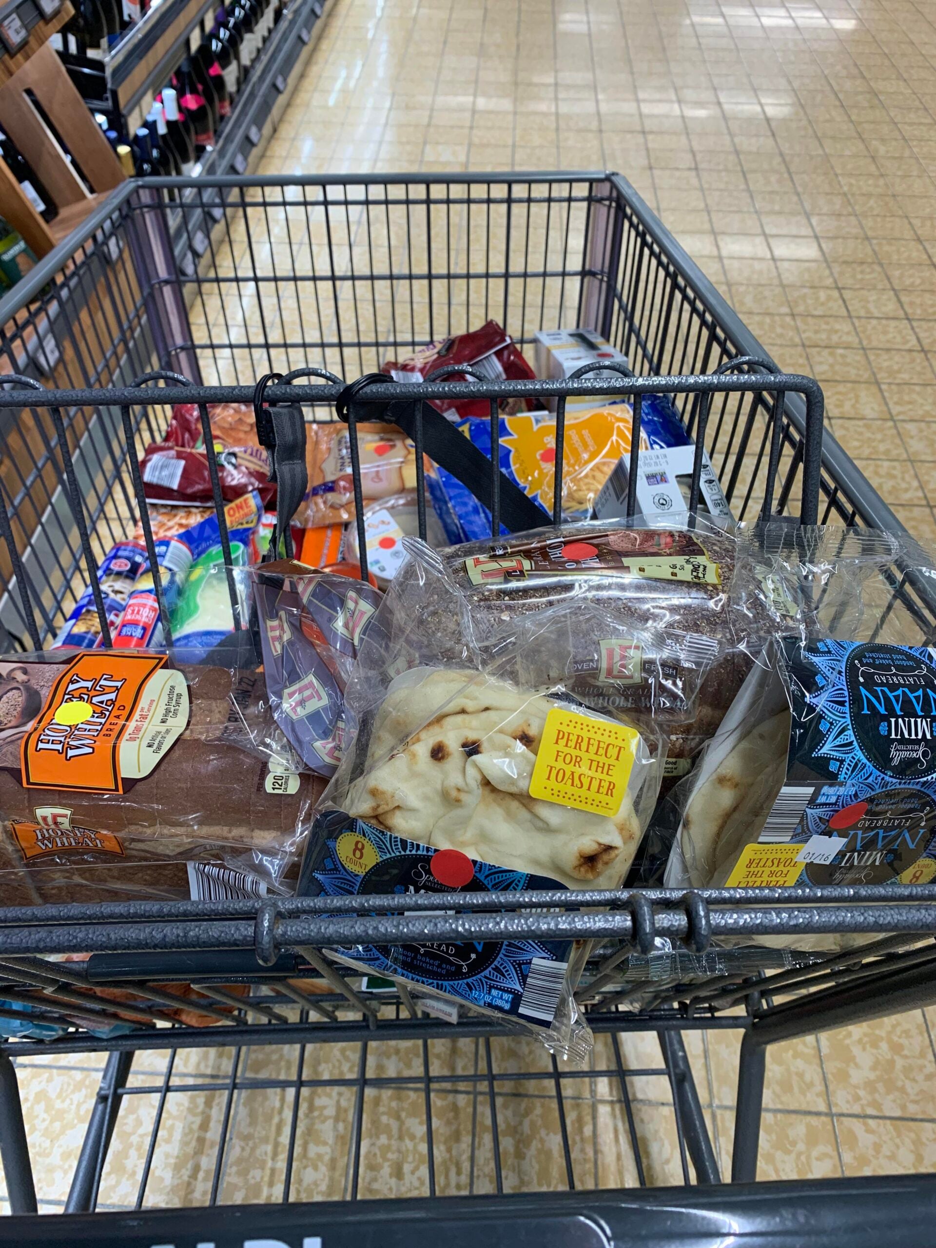 A shopping cart filled with various groceries, including bread, naan, cheese, and packaged foods, is seen in a grocery store aisle with a tiled floor.
