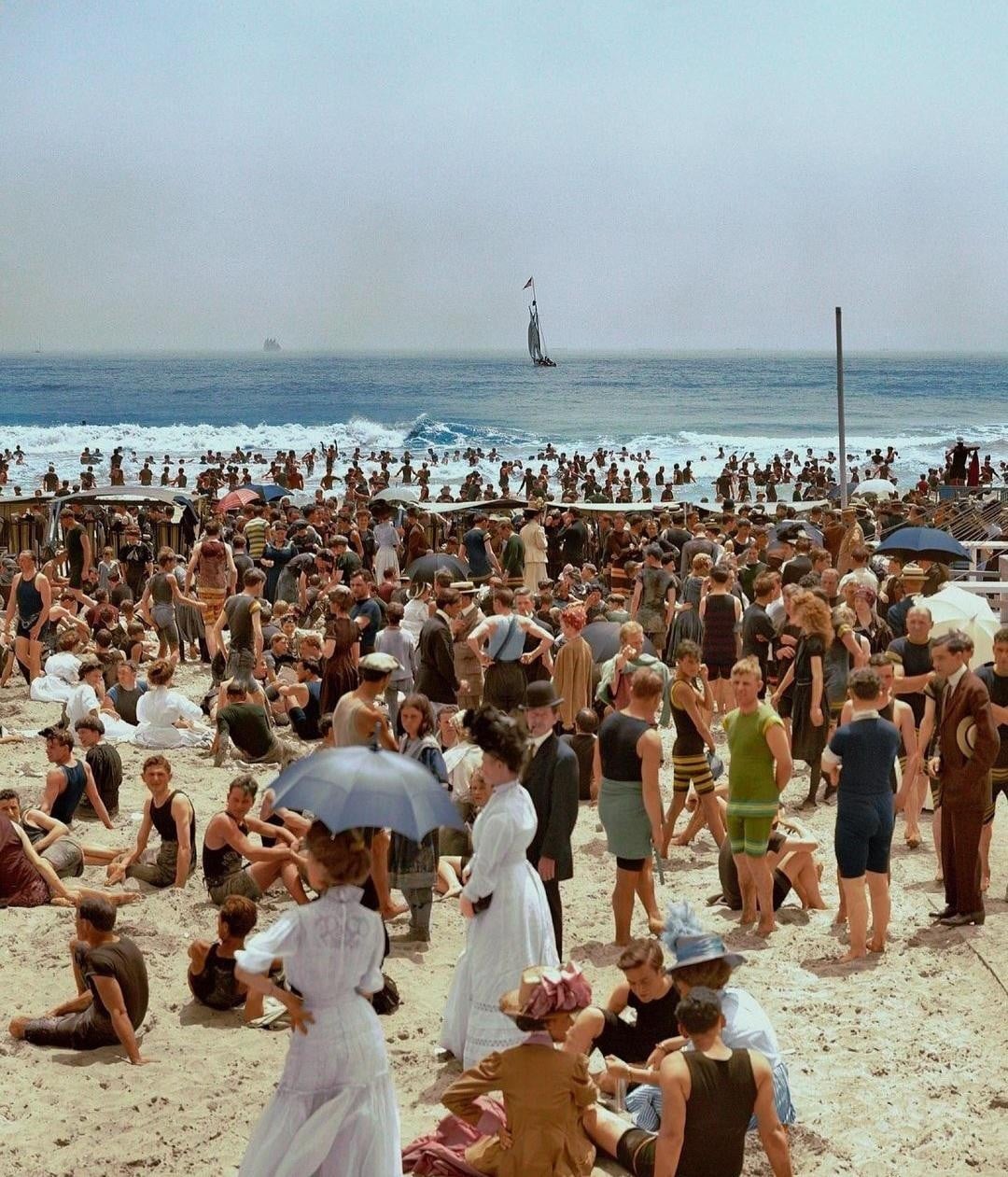 A crowded beach scene from the early 1900s, with people in vintage swimsuits and hats. Some sit or stand on the sand, while others swim in the ocean. A sailboat is visible on the water under a clear sky.