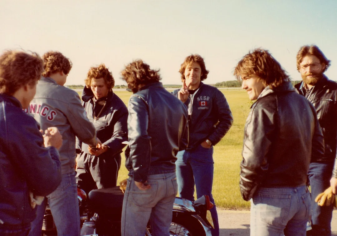 Seven men wearing jackets stand around a motorcycle on a sunny day in a grassy open field. Some are talking, one is smoking, and another is making a peace sign while smiling at the camera.