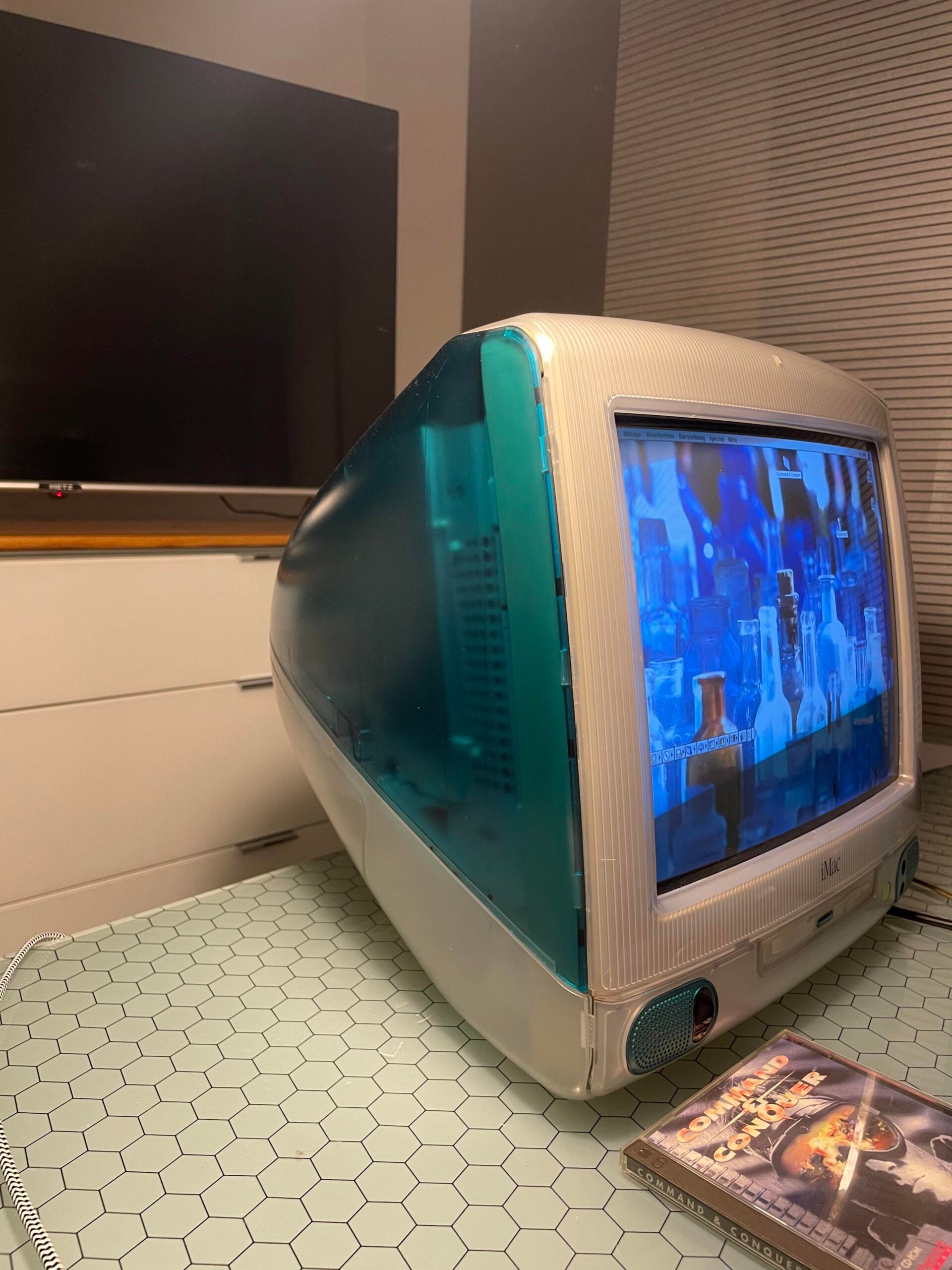 A teal and white iMac G3 computer sits on a hexagonal-tiled table, displaying a blue-tinted screen. A DVD copy of “Men in Black II” is next to it. A modern flat-screen TV is in the background.