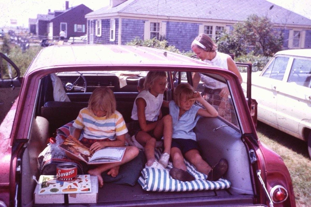 Three children sit in the back of an open station wagon reading and playing with snacks and games, while a woman stands outside by the car door. A house and another car are visible in the background.