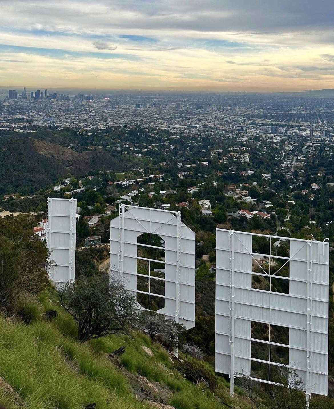 View from behind the iconic Hollywood sign overlooking Los Angeles, with the cityscape and skyline visible under a cloudy sky. Green hills and homes appear in the foreground.