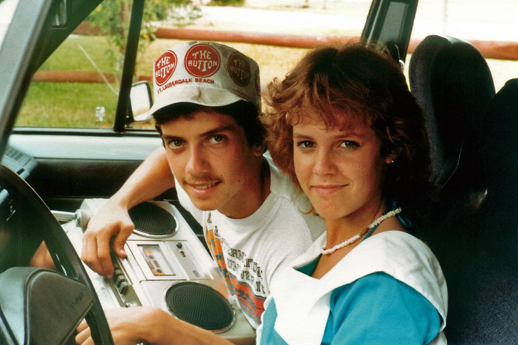 Two young people sit in a car; the man holds a boombox and wears a cap with “The Button” badges. The woman, in a blue top and white necklace, smiles at the camera.