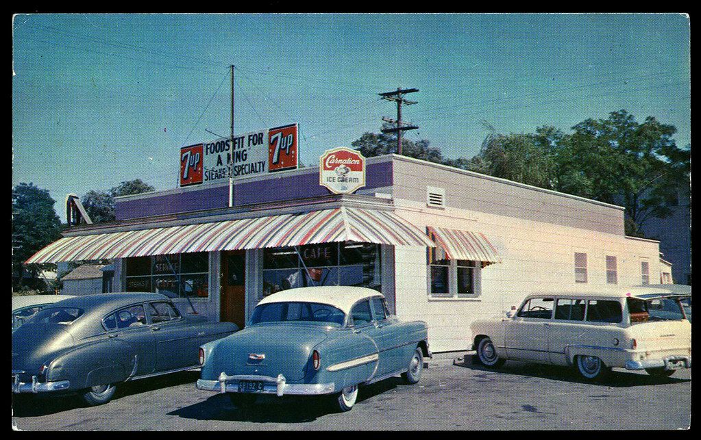 Vintage diner with striped awning, 7UP signs, and three classic 1950s cars parked outside. A cafe sign is visible above the entrance. Trees and power lines are in the background.
