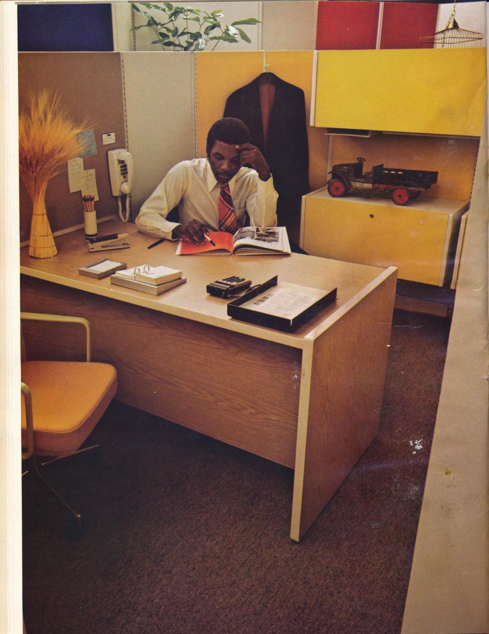 A man in a shirt and tie sits at a wooden office desk reading a magazine. The office has yellow cabinets, books, a rotary phone, a wheat decoration, and a model train on the shelf behind him.