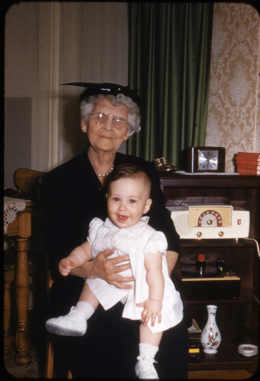 An elderly woman in glasses and a black dress holds a smiling baby in a white outfit. They sit indoors beside a vintage radio and clock, with green curtains and patterned wallpaper in the background.