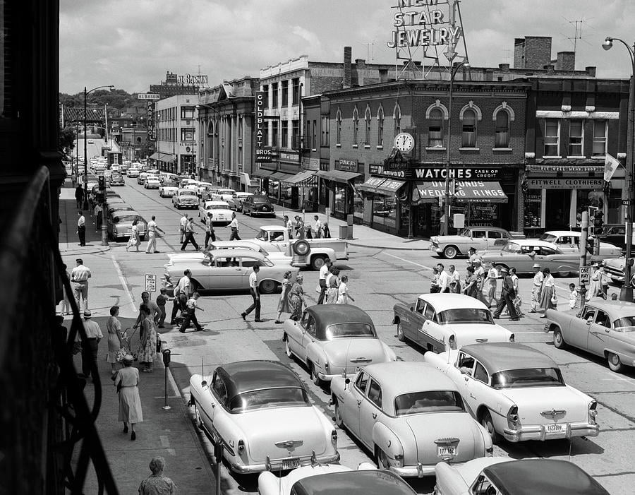 A busy 1950s city street with vintage cars parked and driving, and many people crossing the intersection. Storefronts with signs, including one for Star Jewelry, line the sidewalks under a partly cloudy sky.