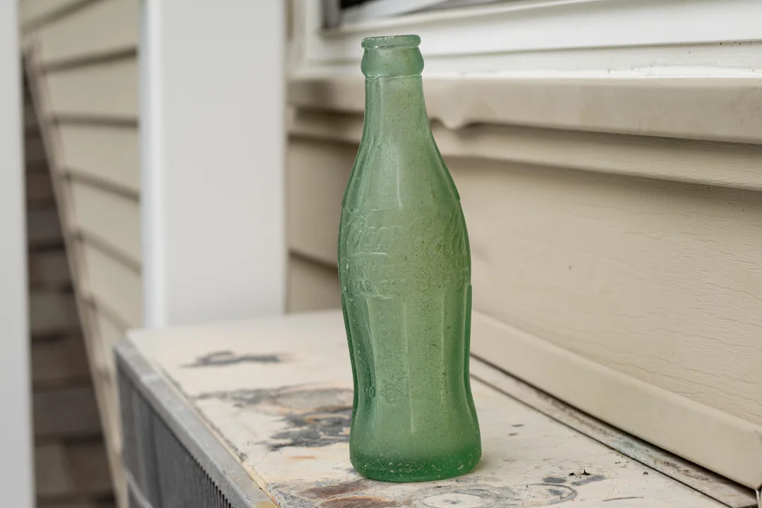 A vintage green glass Coca-Cola bottle sits on a dusty, weathered surface outdoors, near beige siding and a window ledge.