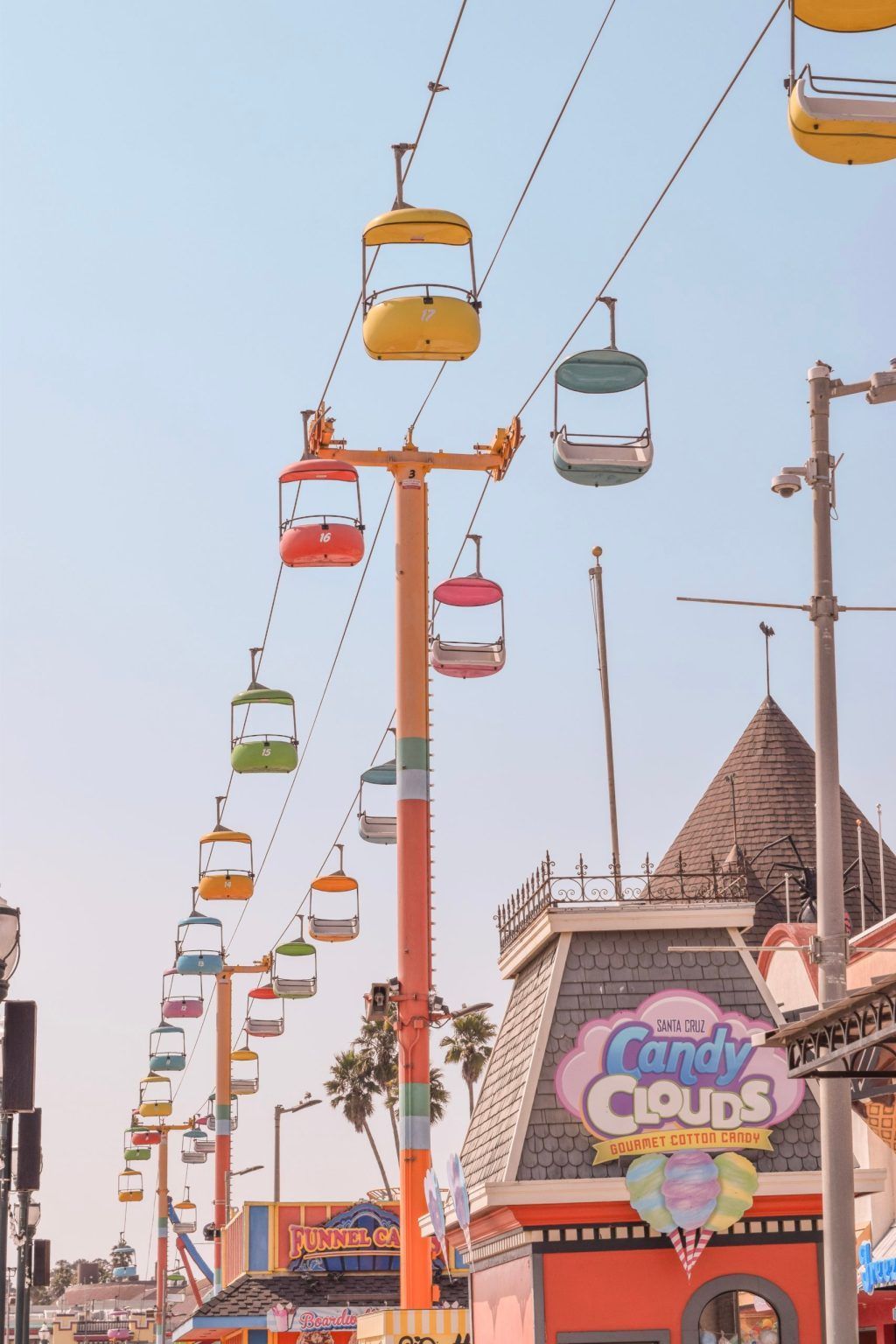 Colorful sky ride gondolas travel above a boardwalk with pastel buildings, including one with a sign reading "Candy Cups," under a clear blue sky. Palm trees and amusement park signs are visible in the background.
