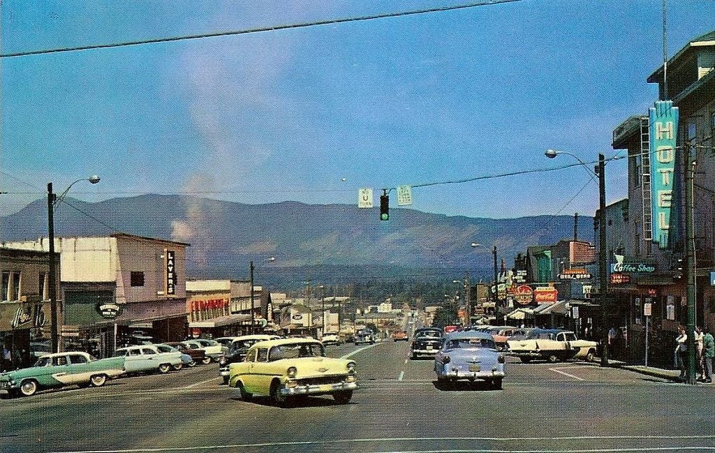 A colorful 1950s street scene shows classic cars driving and parked along a busy downtown, with neon hotel and cafe signs, storefronts, and distant mountains under a blue sky.