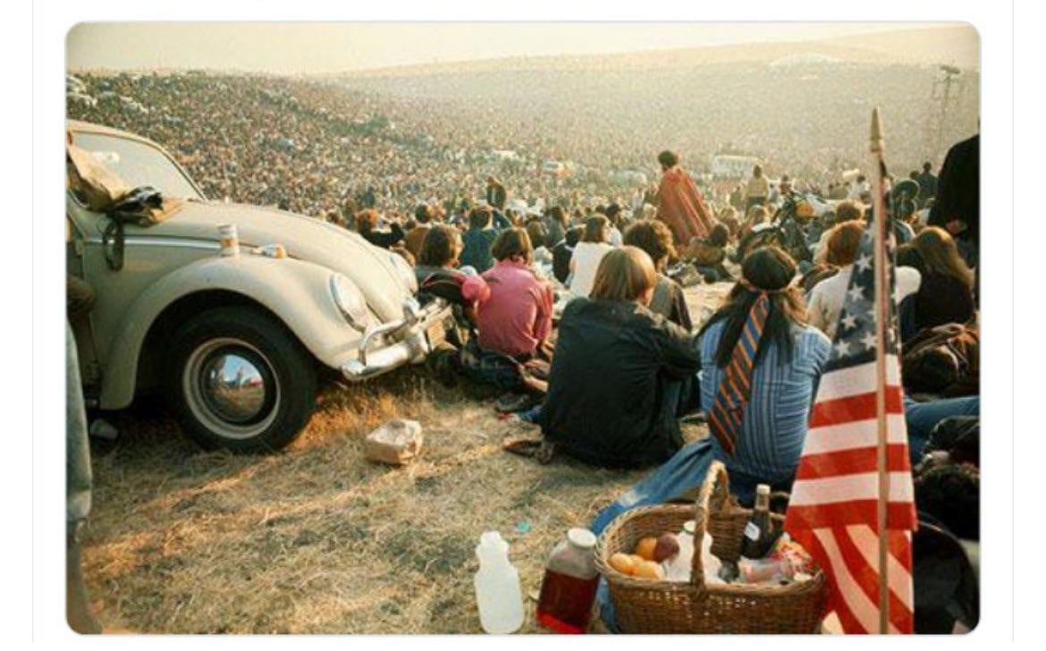 A large crowd sits on a grassy hillside by a Volkswagen Beetle, with a picnic basket and an American flag in the foreground, at an outdoor event or festival under hazy skies.