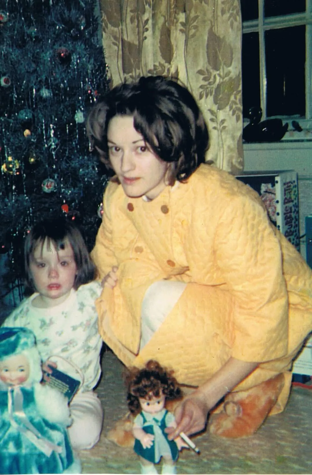 A woman in a yellow robe poses with a young child in pajamas in front of a silver Christmas tree, with dolls on the floor and wrapped presents nearby.