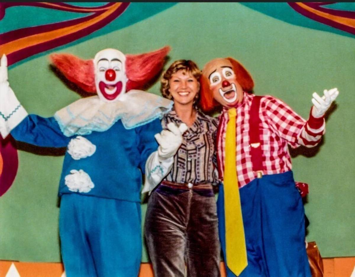 A woman stands smiling between two clowns in colorful costumes, one in blue with red hair and the other in a red checkered shirt with yellow tie, against a green and rainbow backdrop.