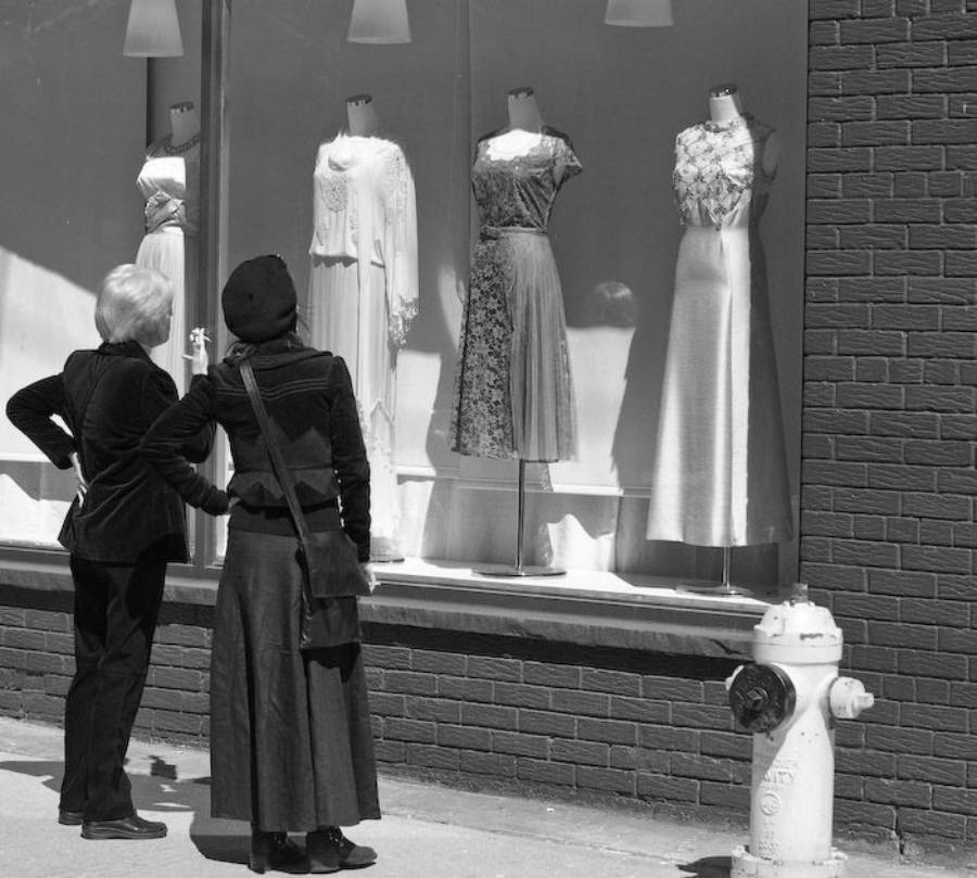 Two women stand on a sidewalk, looking at elegant dresses displayed on mannequins in a store window. A fire hydrant is visible in the foreground. The scene is in black and white.